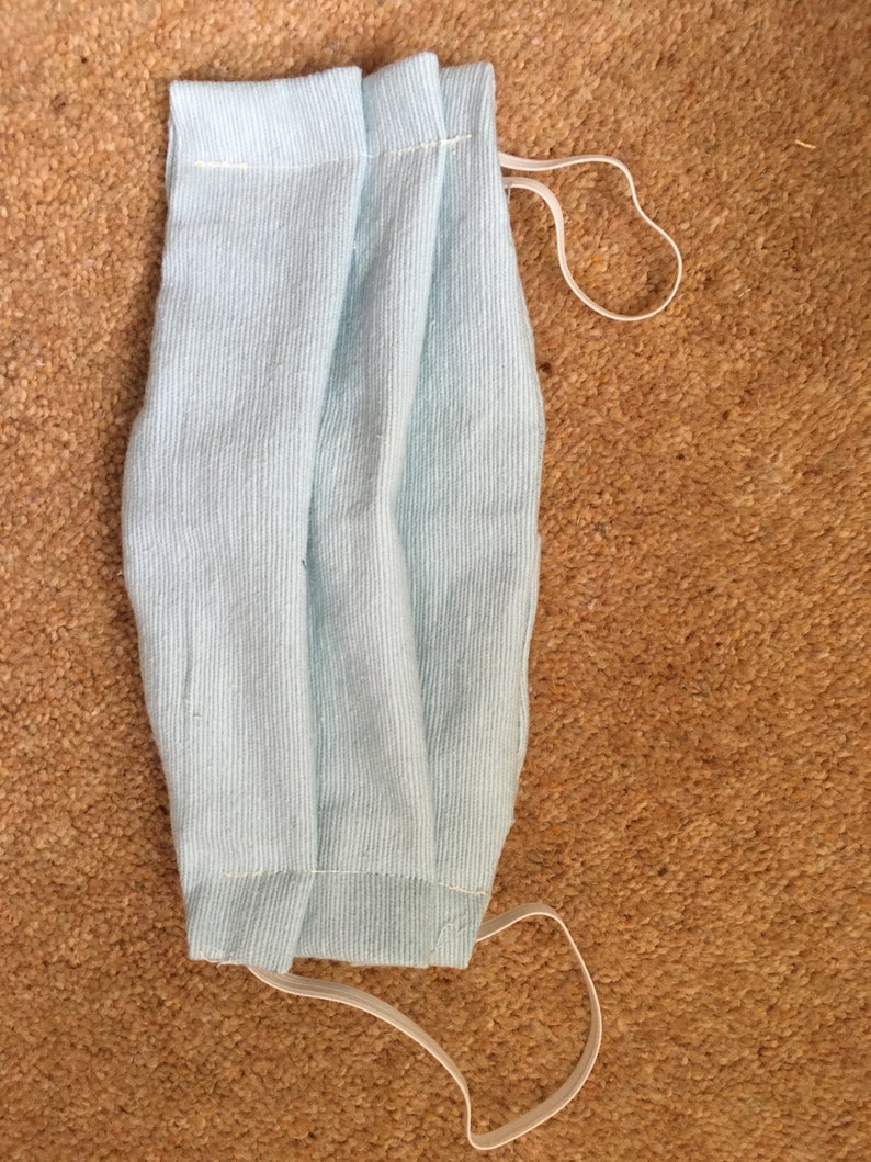 May include: A light blue fabric face mask with white elastic ear loops. The mask is folded in half and has a pleated design.
