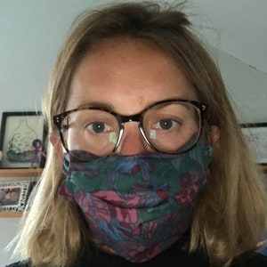 May include: A person wearing a patterned face mask and brown framed glasses. The mask is a dark blue, green, and pink floral pattern.