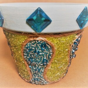 Puede incluir: Un pequeño macetero de cerámica blanca decorado con pedrería azul y adornado con cuentas amarillas y azules.