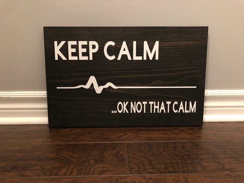 Puede incluir: Un letrero de madera marr&oacute;n oscuro con texto blanco que dice "KEEP CALM...OK NOT THAT CALM". Una l&iacute;nea EKG blanca est&aacute; por encima del texto.