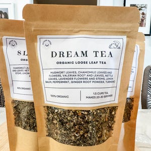 Pode incluir: Um saco de papel castanho com uma janela transparente que mostra uma mistura de ervas e flores secas. O saco é etiquetado como "Dream Tea Organic Loose Leaf Tea" e lista os ingredientes: folhas de artemísia, folhas e flores de camomila, raiz e folhas de valeriana, folhas de urtiga, flores e caules de lavanda, bálsamo de limão, hortelã-pimenta, pó de raiz de gengibre, açafrão. O saco também indica "100% orgânico" e "1,5 chávenas de chá, faz 20-30 porções."
