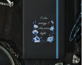 Könnte beinhalten: Schwarzes Journal mit einem blauen Band-Lesezeichen. Der Einband zeigt ein florales Design mit blauen Blumen und dem Text "Ich bin Energie. Ich bin reines Licht."
