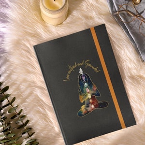 May include: A black journal with the text "I am centered and Grounded" in gold script. The journal features a colorful illustration of a person in a seated yoga pose. A gold-colored band runs vertically along the right side.