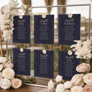 May include: A clear acrylic wedding seating chart frame featuring six navy blue cards labeled Table One through Table Six with white text and gold star accents. Each card is secured with a wax seal and the frame is decorated with white floral arrangements.