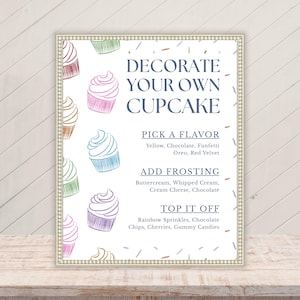 以下が含まれることがあります： ビーズの縁取りが施された長方形の看板で、DECORATE YOUR OWN CUPCAKE、PICK A FLAVOR、ADD FROSTING、TOP IT OFFという文字が書かれています。白い背景には、カップケーキとスプリンクルのカラフルな線画が描かれています。