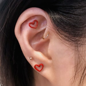 May include: Close-up of an ear with multiple piercings. Two red heart-shaped earrings are visible, along with a small gold stud and a gold hoop earring. The skin tone is fair.