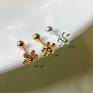 May include: Three flower-shaped stud earrings in rose gold, gold, and silver. Each earring has a small ball on one end and a flower design on the other. The words "rose gold", "gold", and "silver" are written next to each earring.