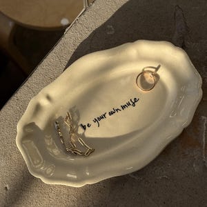 May include: An oval-shaped cream-coloured ceramic trinket dish with scalloped edges. The centre features the black handwritten text 'be your own muse'. A gold-toned metal ring and a gold-toned metal chain link bracelet rest on the dish surface.
