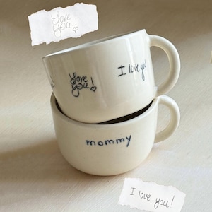 Puede incluir: Dos tazas de cerámica blanquecina apiladas. La taza superior tiene escrito "I love you" y "Love you!" con tinta negra. La taza inferior dice "mommy". Dos notas de papel rasgadas dicen "Love you!" e "I love you!"