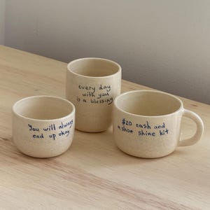 May include: Three beige ceramic mugs with handwritten messages. The mugs are on a light brown wooden surface. The messages on the mugs are: "You will always end up okay", "Every day with you is a blessing", and "$20 cash and a shoe shine kit".