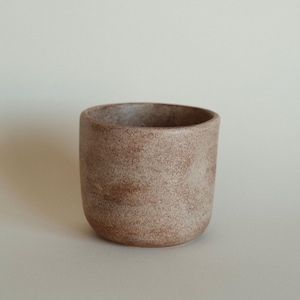 May include: A small, round, ceramic container with a speckled, earthy brown finish. The vessel has a smooth, rounded base and a wide opening. The neutral color and simple design suggest a minimalist aesthetic, suitable for holding small items or as a decorative piece.