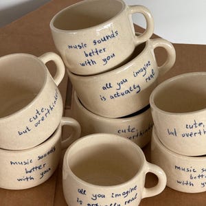 May include: A set of six beige ceramic mugs with blue handwritten text. The text on the mugs reads "music sounds better with you", "all you imagine is actually real", and "cute, but overthinker".