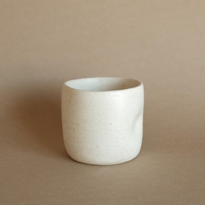 Può includere: Una piccola tazza in ceramica bianco sporco, di forma arrotondata e superficie leggermente ruvida. La tazza ha un interno liscio e un design semplice e minimalista. La tazza viene probabilmente utilizzata per bere bevande.