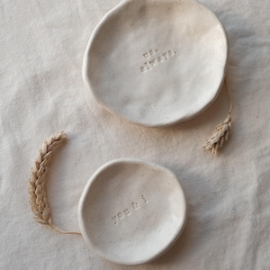 Puede incluir: Dos pequeños cuencos de cerámica blanquecina con las frases "us. always." y "you & i" grabadas en relieve. Los cuencos tienen forma irregular y están sobre una tela de color neutro. Una espiga de trigo seca se coloca cerca de los cuencos.