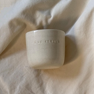 Puede incluir: Una taza de cerámica blanquecina moteada con la frase "I am enough" grabada en la parte delantera. La taza tiene forma cilíndrica y está sobre una tela de tono neutro. La taza es de tamaño pequeño.