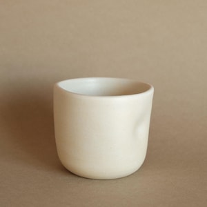 May include: A small, off-white ceramic cup with a smooth, matte finish and a rounded, cylindrical shape. The vessel features a subtle, singular indentation on its side, creating a soft, organic contour along the exterior wall of the cup.