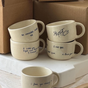 Op de afbeelding: Vijf crèmekleurige keramische mokken met gespikkelde texturen en zwarte handgeschreven tekst. De mokken bevatten zinnen zoals "Too committed, forever", "Stephen", "I love you Right, Now Right, Forever -J", "I love you" en "i love you always".