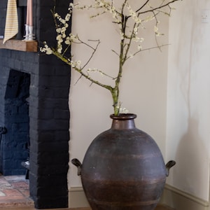 May include: A large, dark brown, ceramic vase with two handles sits in the corner of a room. The vase is filled with a branch of white blossoms.
