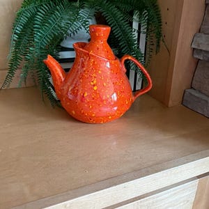 May include: A bright orange ceramic teapot featuring a speckled yellow and red paint splatter pattern. The teapot has a rounded body, a curved handle, a pointed spout, and a matching lid with a small knob handle on top for easy removal.