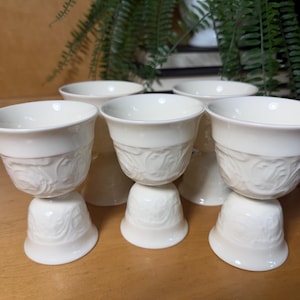 May include: A set of five off-white ceramic egg cups featuring a raised, embossed scrollwork pattern around the bowl and base. Each cup has a wide, flared rim and a sturdy pedestal foot, providing a consistent look for serving soft-boiled eggs.