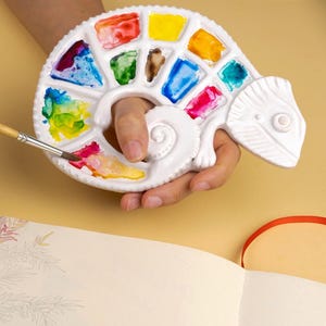 Może przedstawiać: Biała ceramiczna paleta w kształcie kameleona wypełniona kolorowymi farbami akwarelowymi. Pędzel trzymany nad paletą, a na pierwszym planie pusty notatnik z czerwoną obw&oacute;dką. Artykuły plastyczne.