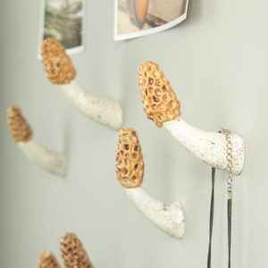 May include: Four white ceramic mushroom-shaped hooks with brown tops are mounted on a wall. A brown leather bag hangs from one hook, and a white beaded necklace hangs from another.