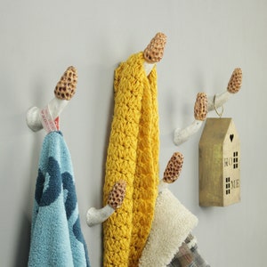 May include: White ceramic mushroom-shaped hooks with a brown top mounted on a gray wall. A blue towel, a yellow crocheted scarf, and a brown plaid jacket are hanging on the hooks. A small gold house with the word "HOME" on it is hanging on the right side of the image.