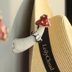 Puede incluir: Ganchos de cerámica blanca en forma de hongo con detalles rojos y blancos. Un sombrero de paja con una cinta negra que dice "LeilyCloud" está colgado en uno de los ganchos.
