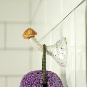 May include: A white ceramic mushroom-shaped hook with brown accents mounted on a white tiled wall. A purple and green scrub brush hangs from the hook.