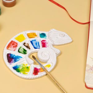 Może przedstawiać: Biała ceramiczna paleta w kształcie kameleona wypełniona kolorowymi farbami akwarelowymi. Pędzel spoczywa na palecie. Widoczne są r&oacute;wnież słoiczki z farbą i szkicownik, co sugeruje motyw artystyczny lub rzemieślniczy.