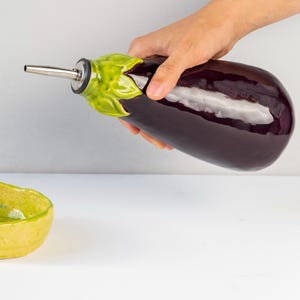 Può includere: Una bottiglia di olio in ceramica a forma di melanzana con beccuccio in acciaio inossidabile, tenuta in mano. La bottiglia è viola scuro con dettagli di foglie verdi. Una piccola ciotola di ceramica gialla è nelle vicinanze.