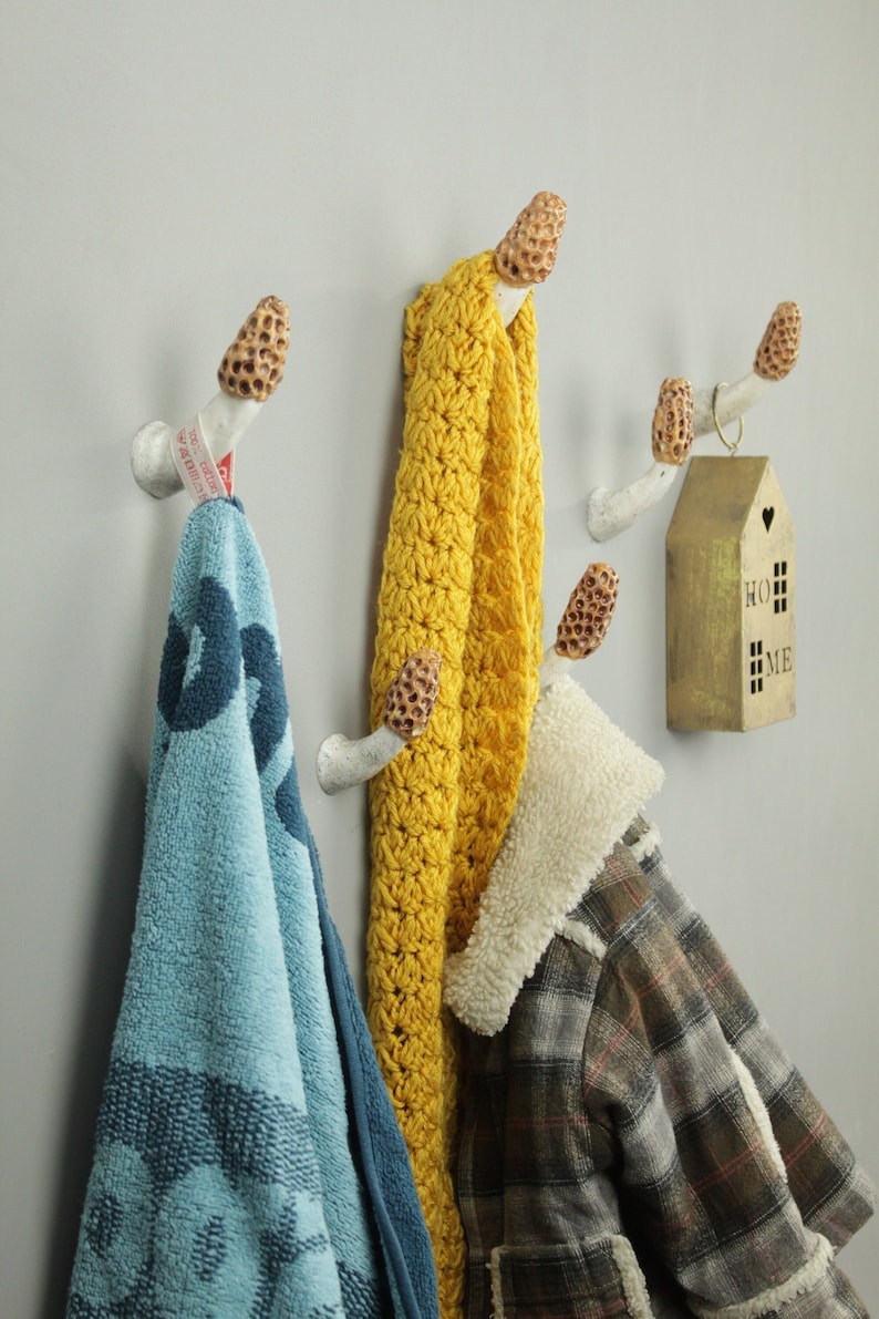May include: White ceramic mushroom-shaped hooks with a brown top mounted on a gray wall. A blue towel, a yellow crocheted scarf, and a brown plaid jacket are hanging on the hooks. A small gold house with the word "HOME" on it is hanging on the right side of the image.