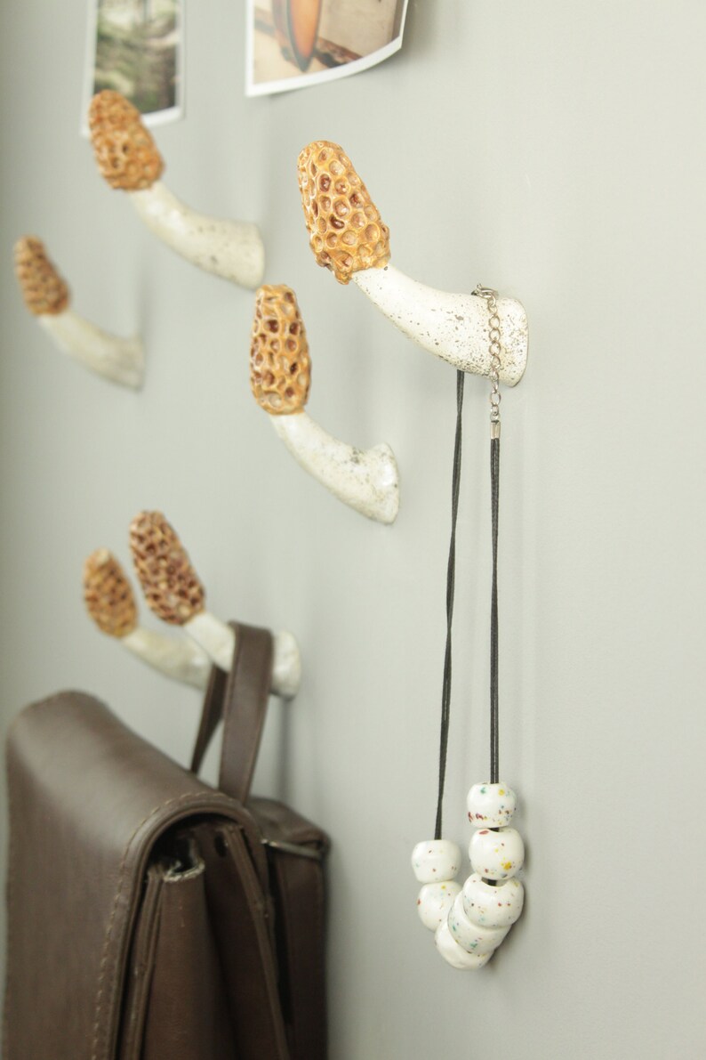 May include: Four white ceramic mushroom-shaped hooks with brown tops are mounted on a wall. A brown leather bag hangs from one hook, and a white beaded necklace hangs from another.