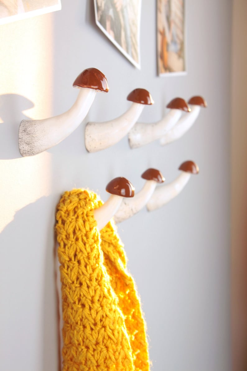 May include: White ceramic mushroom-shaped hooks with brown tops mounted on a wall. A yellow crocheted scarf is hanging on one of the hooks.