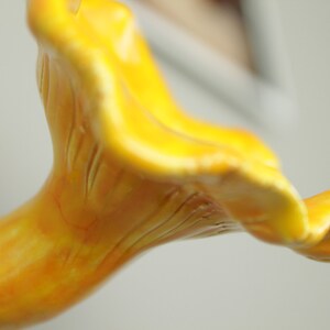 May include: A close-up of a yellow ceramic mushroom sculpture with a detailed, textured surface. The mushroom is shaped like a chanterelle with a wavy cap and a long, slender stem.