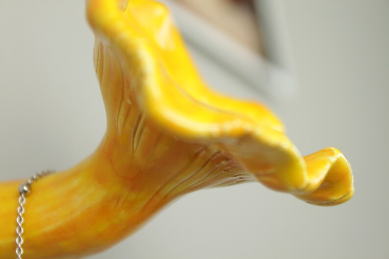 May include: A close-up of a yellow ceramic mushroom sculpture with a detailed, textured surface. The mushroom is shaped like a chanterelle with a wavy cap and a long, slender stem.
