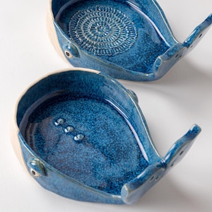 May include: Two ceramic whale-shaped dishes with a glossy blue glaze. The dishes have a white rim and tail, with a textured design in the center of one. The other dish has three small, raised dots. These decorative dishes are ideal for holding small items.