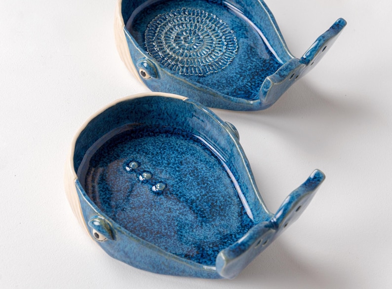 May include: Two ceramic whale-shaped dishes with a glossy blue glaze. The dishes have a white rim and tail, with a textured design in the center of one. The other dish has three small, raised dots. These decorative dishes are ideal for holding small items.