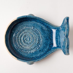 May include: A blue ceramic fish-shaped dish with a textured interior. The dish has a white rim and a tail fin. The interior features a spiral pattern. The dish is on a white surface.