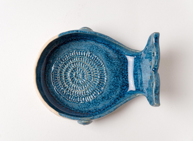 May include: A blue ceramic fish-shaped dish with a textured interior. The dish has a white rim and a tail fin. The interior features a spiral pattern. The dish is on a white surface.