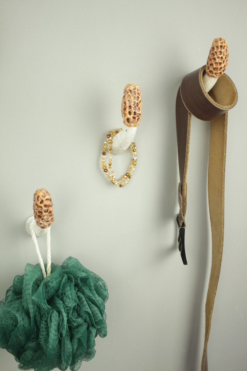 May include: Three white ceramic hooks shaped like mushrooms with brown tops. A green bath pouf hangs from one hook, a brown leather belt hangs from another, and a gold beaded necklace hangs from the third.
