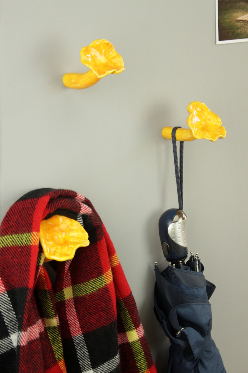 May include: Two yellow ceramic mushroom-shaped hooks mounted on a gray wall. One hook has a red and black plaid scarf hanging from it, and the other has a blue umbrella hanging from it.