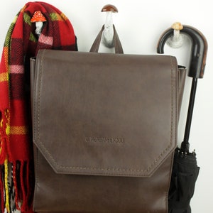 May include: A brown leather backpack with the text "DINGNEANFENDOU" on the front flap. The backpack is hanging on a white hook with a mushroom-shaped top. A red and black plaid scarf is hanging on the hook to the left of the backpack. A black umbrella is hanging on a hook to the right of the backpack.