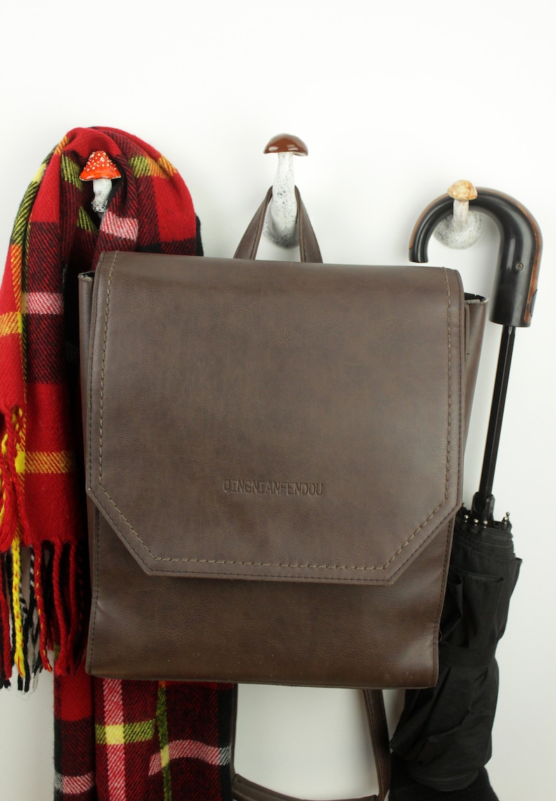 May include: A brown leather backpack with the text "DINGNEANFENDOU" on the front flap. The backpack is hanging on a white hook with a mushroom-shaped top. A red and black plaid scarf is hanging on the hook to the left of the backpack. A black umbrella is hanging on a hook to the right of the backpack.