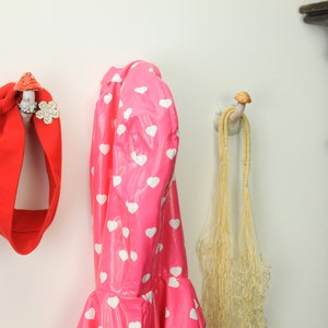 May include: Three hooks on a white wall, each with a different design. The hook on the left has a red headband hanging from it. The middle hook has a pink raincoat with white hearts hanging from it. The hook on the right has a beige net bag hanging from it.