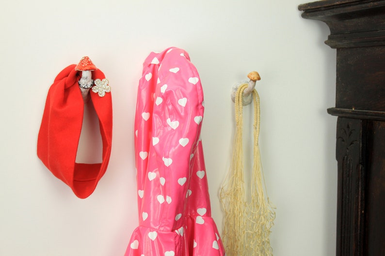 May include: Three hooks on a white wall, each with a different design. The hook on the left has a red headband hanging from it. The middle hook has a pink raincoat with white hearts hanging from it. The hook on the right has a beige net bag hanging from it.