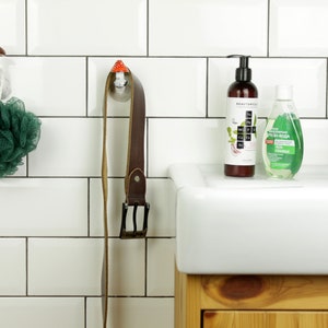 May include: A white tiled bathroom with a white porcelain sink and a wooden cabinet underneath. There are two hooks on the wall, one with a green bath sponge and the other with a brown leather belt. Two bottles of liquid soap are on the counter, one clear and one brown.