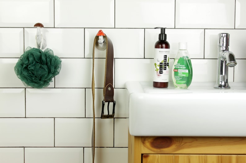 May include: A white tiled bathroom with a white porcelain sink and a wooden cabinet underneath. There are two hooks on the wall, one with a green bath sponge and the other with a brown leather belt. Two bottles of liquid soap are on the counter, one clear and one brown.
