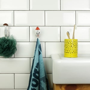 May include: White bathroom tiles with a blue towel hanging on a mushroom-shaped hook. A green bath sponge hangs on a similar hook. A yellow toothbrush holder with two toothbrushes sits on a white countertop next to a chrome faucet.