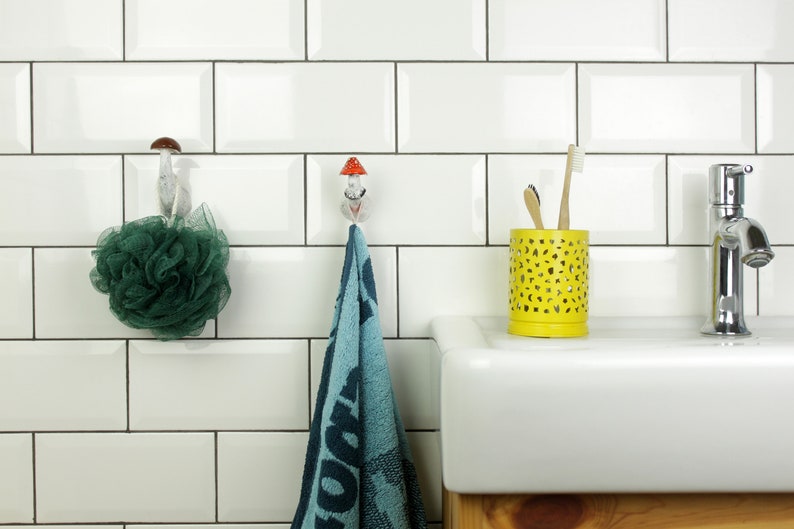 May include: White bathroom tiles with a blue towel hanging on a mushroom-shaped hook. A green bath sponge hangs on a similar hook. A yellow toothbrush holder with two toothbrushes sits on a white countertop next to a chrome faucet.
